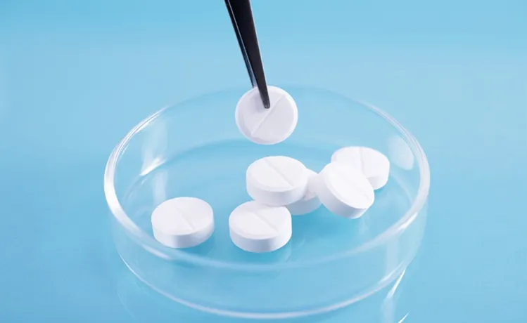 Vignette Actualité - Pincettes ramassant une pilule blanche d'une pile dans un plat en verre dans un laboratoire