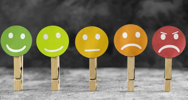 Vignette Actualité : Cinq pinces à linge en bois tiennent des visages colorés exprimant une gamme d’émotions, du bonheur à la colère, illustrant visuellement des sentiments humains.