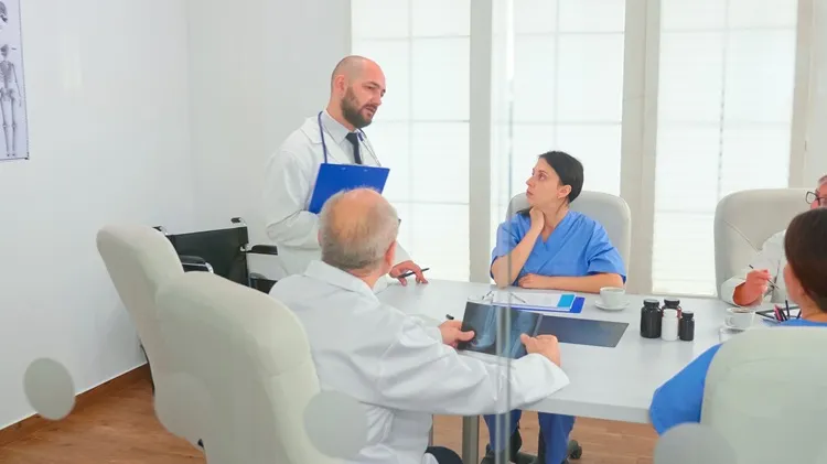 Vignette Actualité : Médecin présentant un diagnostic à des collègues tenant un presse-papiers lors d'un briefing avec des collègues