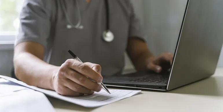 Vignette Actualité : Vue latérale du médecin avec stéthoscope travaillant sur ordinateur portable et écrit sur papier