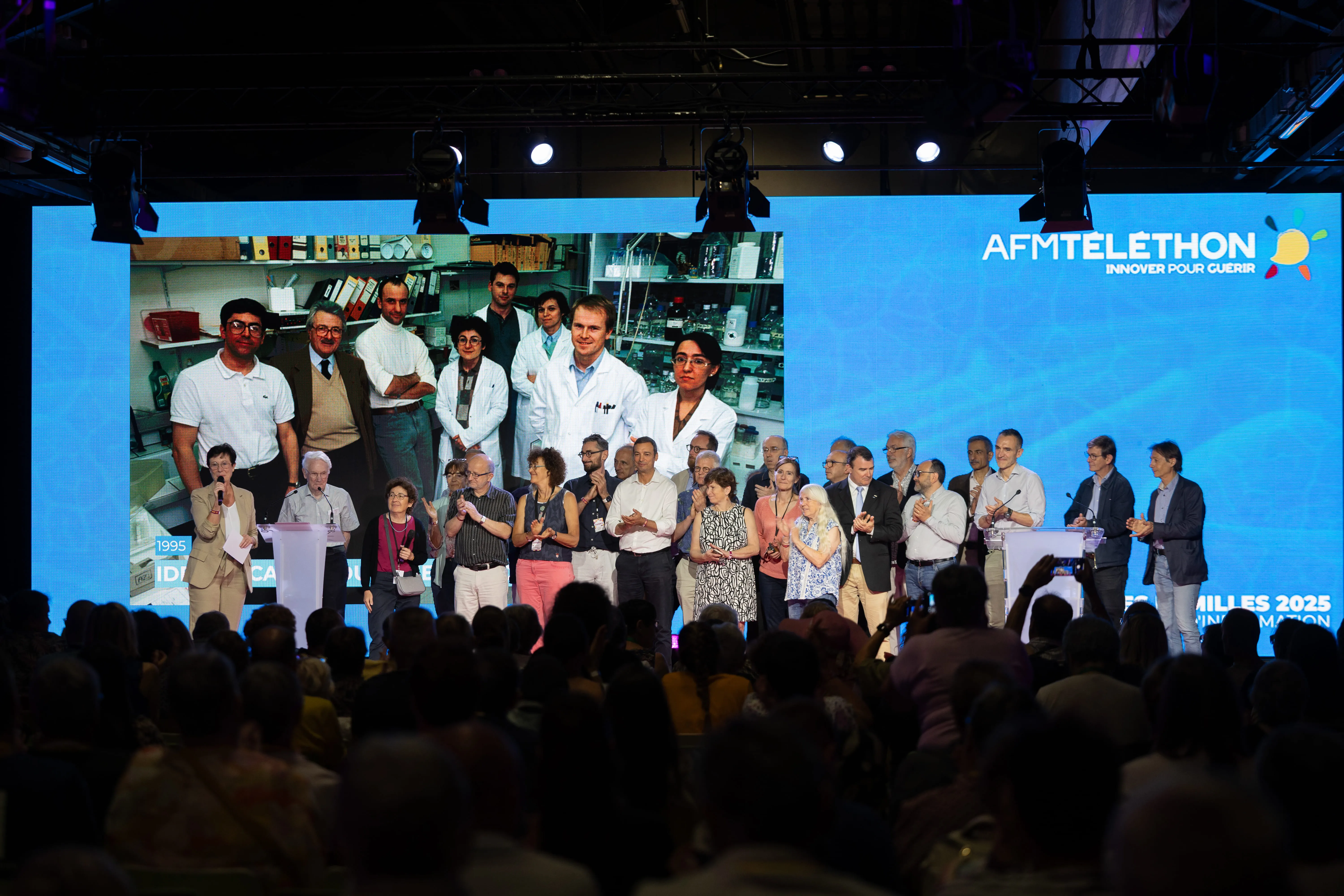 Une vingtaine de personnes, anciens et actuels acteurs de la recherche, rassemblés sur scène pour un hommage collectif, avec en arrière-plan une photo d’archives de 1995. 