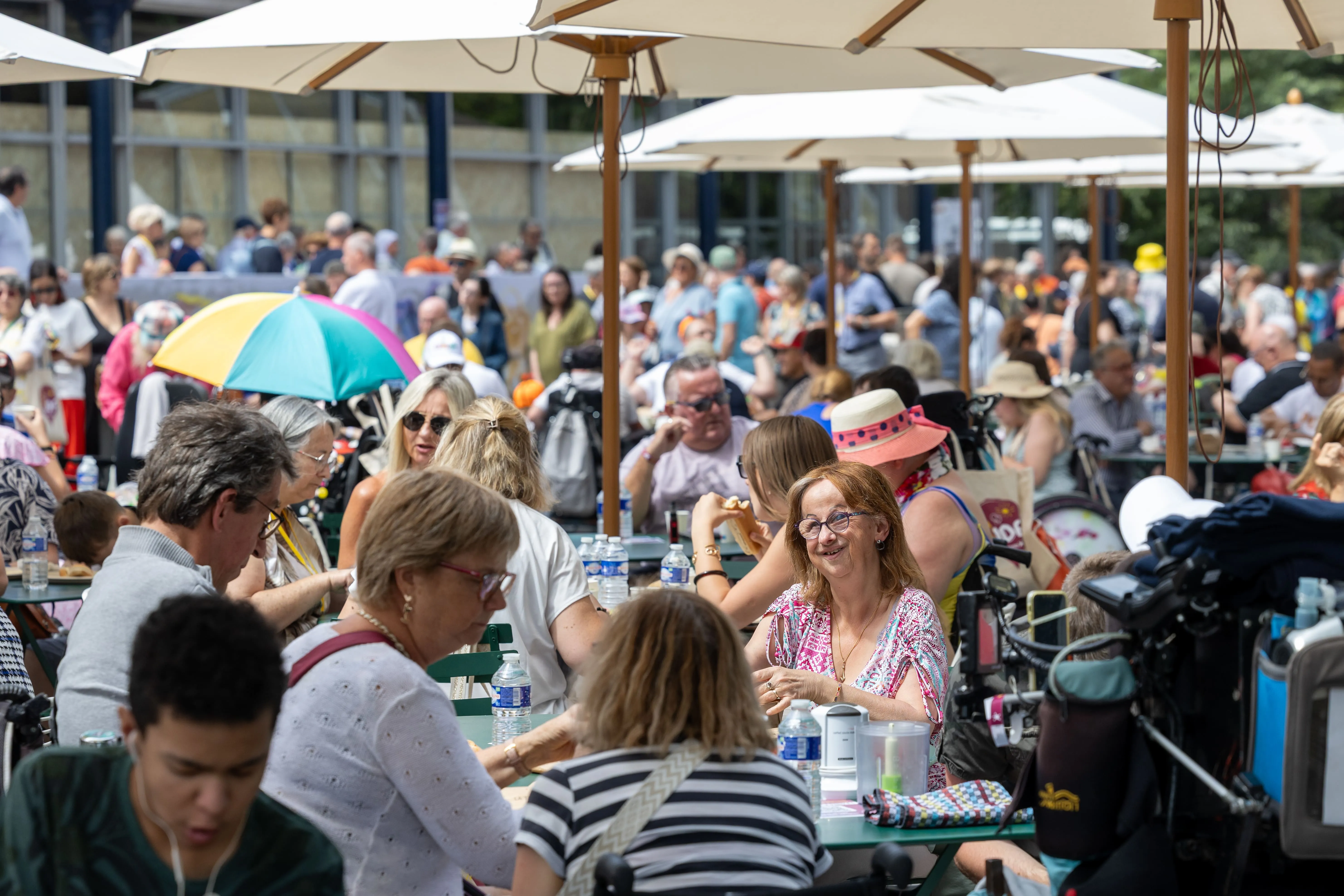 Familles et bénévoles rassemblés autour de grandes tables en extérieur, partageant un moment convivial sous des parasols, dans une ambiance estivale