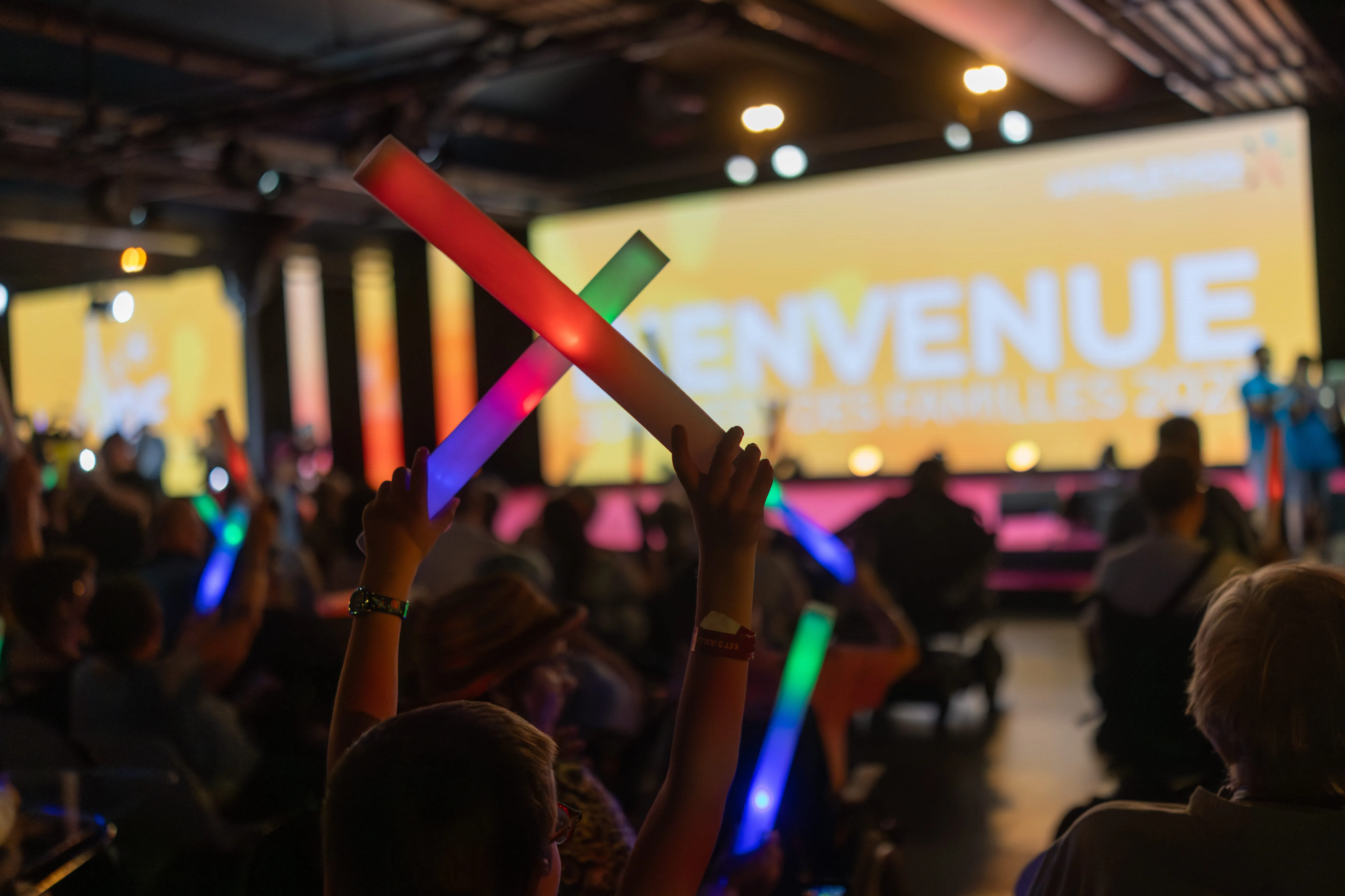 Public enthousiaste agitant des bâtons lumineux multicolores face à une scène aux couleurs vives, où s’affiche le message de bienvenue des Journées des Familles 2025. 