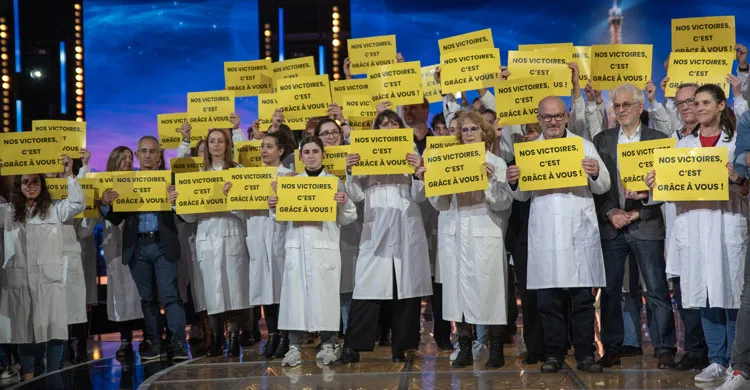 100 chercheurs sur le plateau du Téléthon 2023, tiennent des pancartes "nos victoires c'est grâce à vous"